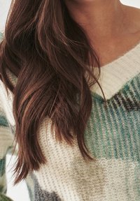 Lange, braune, wellige Haare über einem gestrickten Pullover mit grünen, weißen und grauen Mustern, wobei der Hals- und Schulterbereich sichtbar ist.
