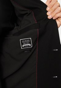 Fodera della giacca nera con etichetta "GUESS", fondata nel 1981, in bianco su sfondo nero; visibile il tag di vestibilità slim; dettaglio in cucitura cremisi.