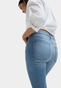 Světle modré džíny s středním pasem, které mají dvě zadní kapsy a hladkou texturu. Nosené s bílou košilí s ohrnutými rukávy.