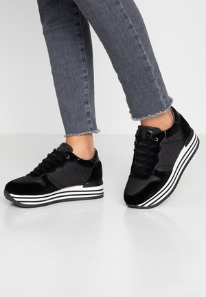 Person, der bærer sorte plateau-sneakers med hvide stribede såler og mørkegrå jeans med frynset kant på en ensfarvet lys gulv.