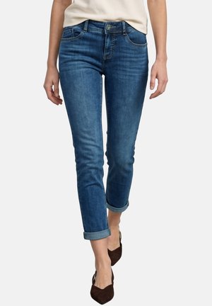 Mujer con vaqueros azul ajustados remangados, camiseta beige y zapatos marrones puntiagudos, caminando sobre un fondo claro y liso.