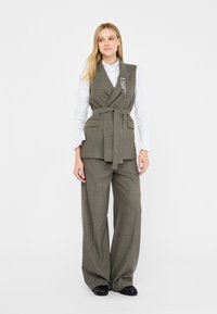 Gilet grigio su misura con design avvolgente, cinture in vita, abbinato a pantaloni larghi coordinati; camicia bianca sotto.