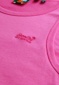 Top de algodón rosa sin mangas con un logo bordado, escote redondeado y detalles de costura visibles. Textura suave con un diseño casual.