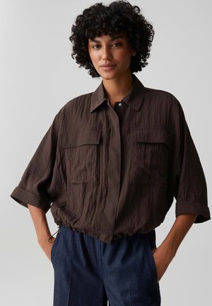 Vrouw met krullend haar die een losse bruine blouse met wijde mouwen en donkerblauwe broek draagt, staand met handen in de zakken tegen een effen achtergrond.