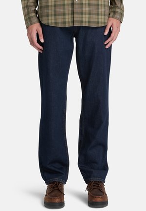 Mann trägt dunkelblaue Jeans, braune Schnürschuhe und ein grünes kariertes Hemd mit langen Ärmeln, steht mit entspannten Händen an den Seiten.