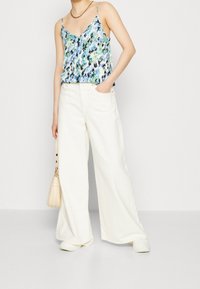 Camisole fluide à motifs bleus et verts avec des bretelles fines, associé à un pantalon large blanc et un sac à main crème, porté de manière décontractée.