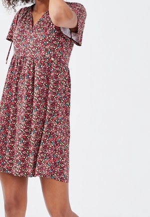 Femme portant une robe à manches courtes à fleurs rouges, roses, bleues et blanches, se tenant devant un fond clair uni.