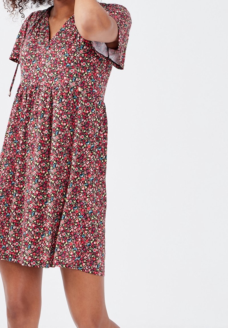 Femme portant une robe à manches courtes à fleurs rouges, roses, bleues et blanches, se tenant devant un fond clair uni.