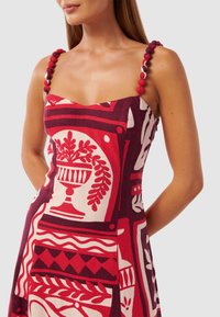 Rode en crèmekleurige jurk met een vierkante halslijn, decoratieve pompon-schouderbanden en een aansluitende silhout. De jurk heeft botanische motieven en geometrische accenten.