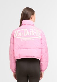 �Ženska z dolgimi rjavimi lasmi, oblečena v krajšega roza puhovka s logotipom "Von Dutch Originals" na hrbtu, v kombinaciji z črnimi leginami.