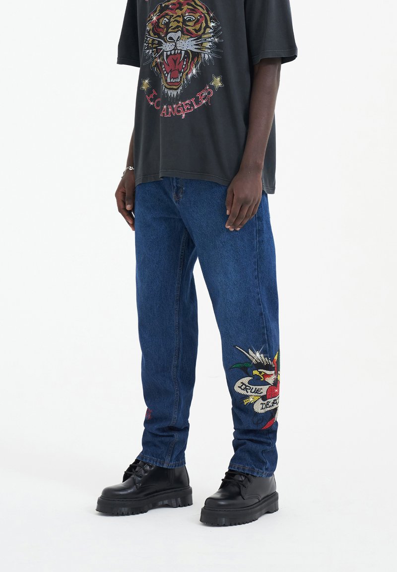 Camiseta gráfica negra con estampado de tigre, combinada con unos vaqueros de mezclilla azules con diseños bordados y botas negras de plataforma.