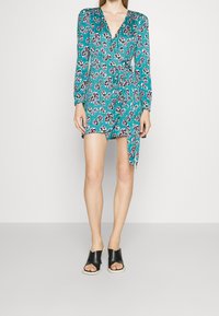 Tee-farbenes Blumenkleid mit langen Ärmeln, V-Ausschnitt und seitlichem Bindegürtel; kombiniert mit schwarz offenen Slippern.