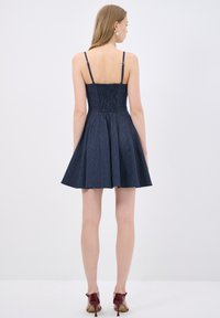 Tummansininen farkkuasu ohuiden hihnojen kanssa, istuva yläosa ja levenevä hame. Teksturoitu, ribatty muotoilu ja rypytetty selkäosa.