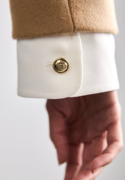 Manche de blazer en laine marron avec un poignet blanc, ornée d'un bouton doré au design floral. Main placée délicatement en dessous du vêtement.
