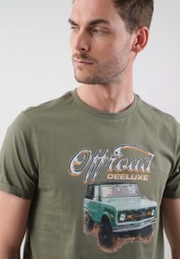 Olivno zelena majica iz bombaža s grafičnim tiskom stare zelene terenske vozila in besedilom "Offroad DEELUXE".