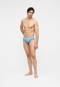 Ανδρικό μοντέλο στέκεται ξυπόλυτο, φοράει ανοιχτό μπλε εσώρουχα Moschino με λογότυπο στη ζώνη, σε απλό λευκό φόντο.