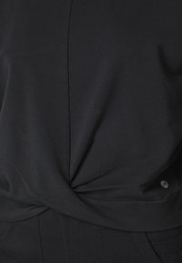 Svart, stickad topp med en tvistad frontdesign och jämn textur. Inkluderar en liten logotags på nedre högra sidan.