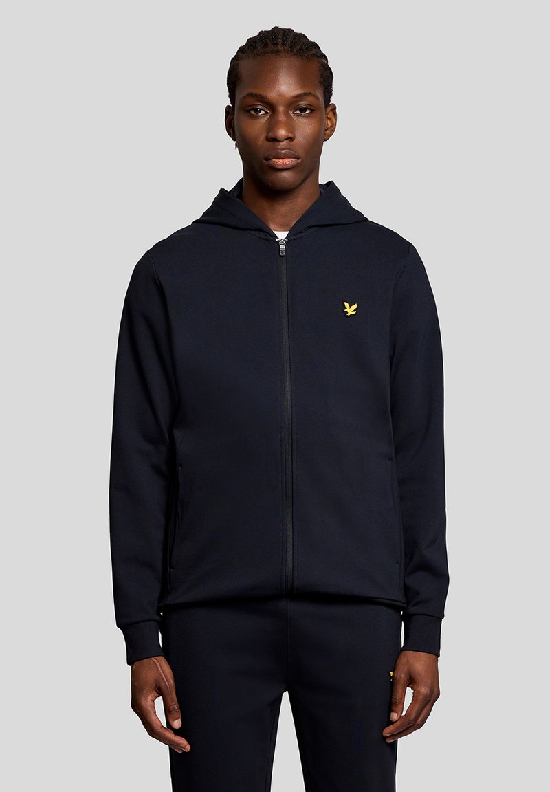 Jeune homme aux cheveux tressés portant un sweat à capuche zippé noir et un pantalon assorti, debout devant un fond gris uni.