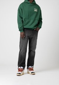 Man med en grön hoodie med vit text, urblekta svarta jeans och vita sneakers med röda och gula snören, stående mot en enfärgad bakgrund.