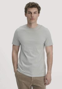 Jonge man met krullend haar, die een lichtgrijze t-shirt met korte mouwen en bruine corduroy broek draagt, met één hand in zijn zak.