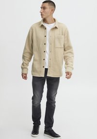 Beige fleece skjortejakke med knappheis foran, en enkelt brystlomme og svarte knapper, kombinert med mørke jeans og svarte joggesko.