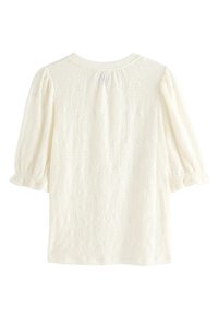 Hvid kortærmet bluse med tekstureret ovalt øjlemønster og elastiske rynkede ærmekanter, vist bagfra på ensfarvet baggrund.