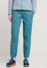 Persoon in teal drawstring broek en kastanjebruine wandelschoenen met blauwe veters, staand met één hand in de zak tegen een effen achtergrond.