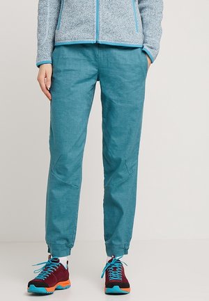 Person wearing teal drawstring pants and maroon hiking shoes with blue laces, standing with one hand in pocket against a plain background.