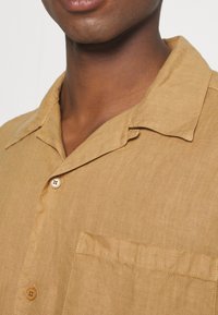 Gros plan d'une personne portant une chemise jaune moutarde à boutons avec un col et une poche poitrine, montrant le bas du visage et le cou.