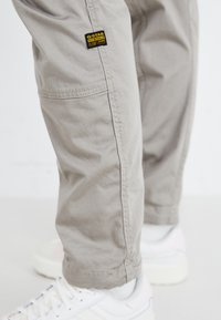 Lys grå cargo-bukser laget av bomull, med sidelommer, synlige sømmer, og et G-Star-merke på låret. Brukt med hvite sneakers.