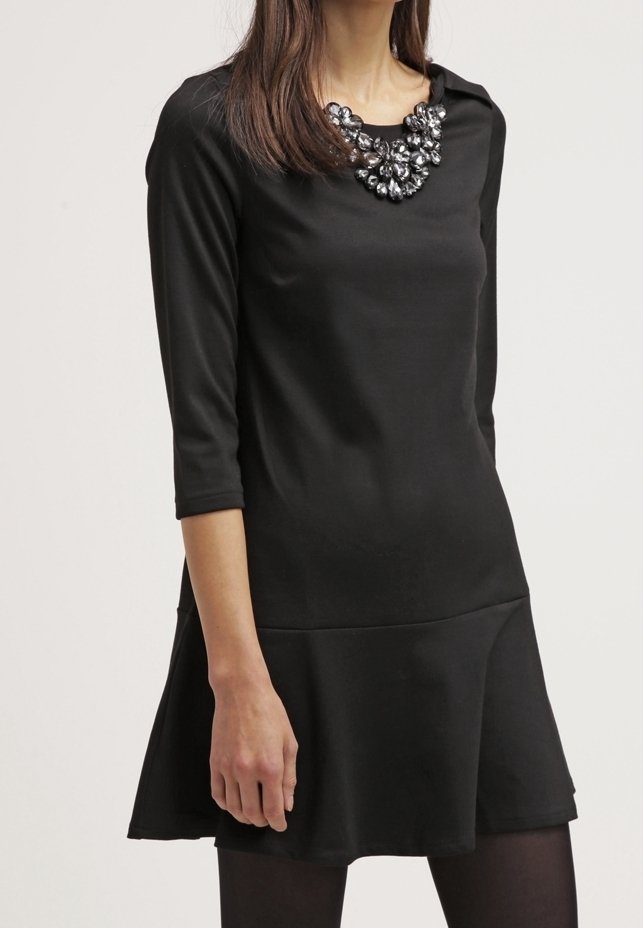 Femme portant une robe noire à manches longues avec une jupe évasée et un grand collier de perles sur un fond clair.