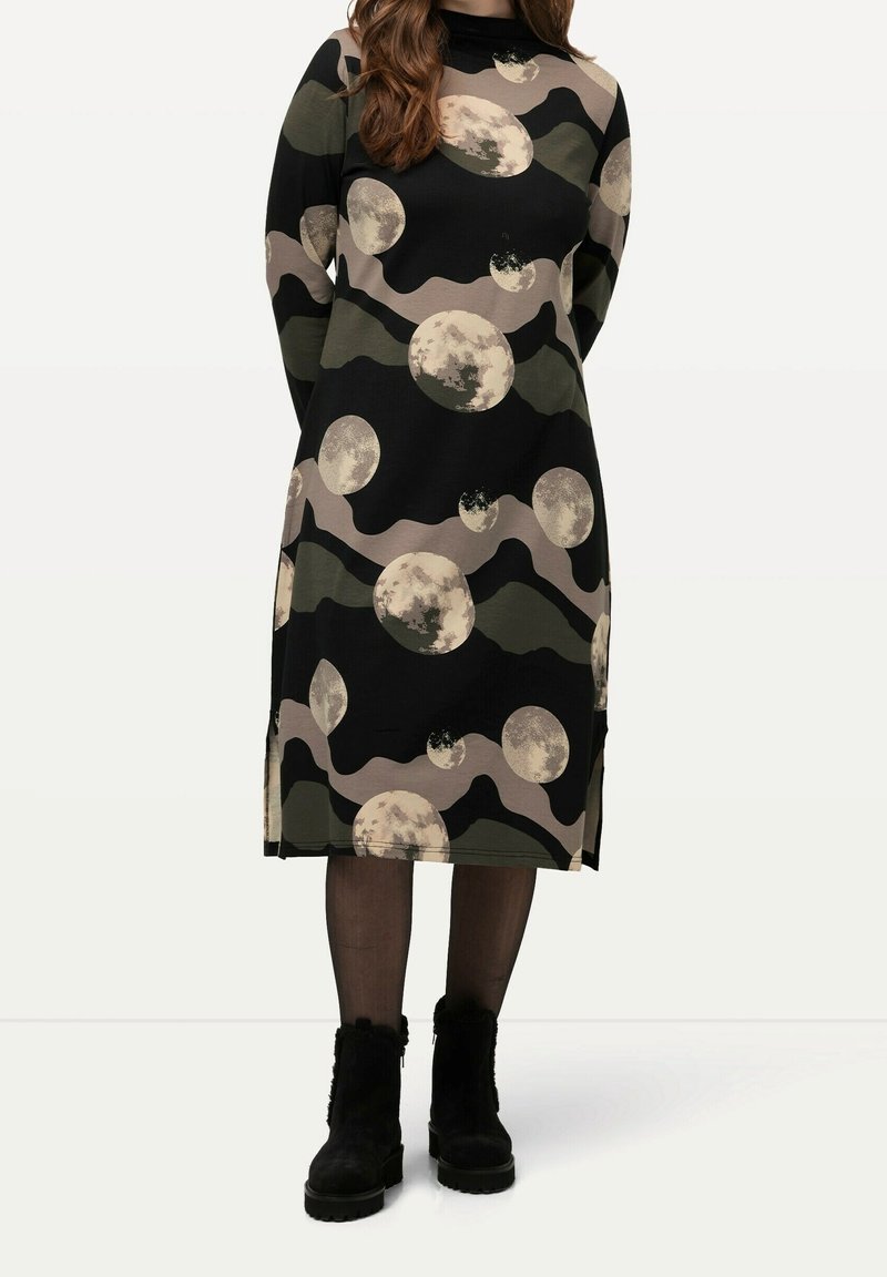 Femme portant une robe midi noire avec des motifs abstraits de lune et de vagues beige et vert olive, des collants noirs transparents et des bottines noires.