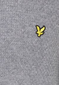 Tissu gris tricoté présentant un motif texturé, avec un petit logo aigle brodé noir et jaune positionné sur le côté gauche.