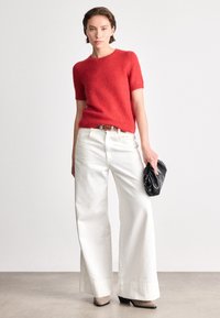 Piros rövid ujjú kötött pulóver, kerek nyakkivágással, magas derekú fehér, bő szárajú nadrággal párosítva. Fekete klu csomagolást tart a kezében.