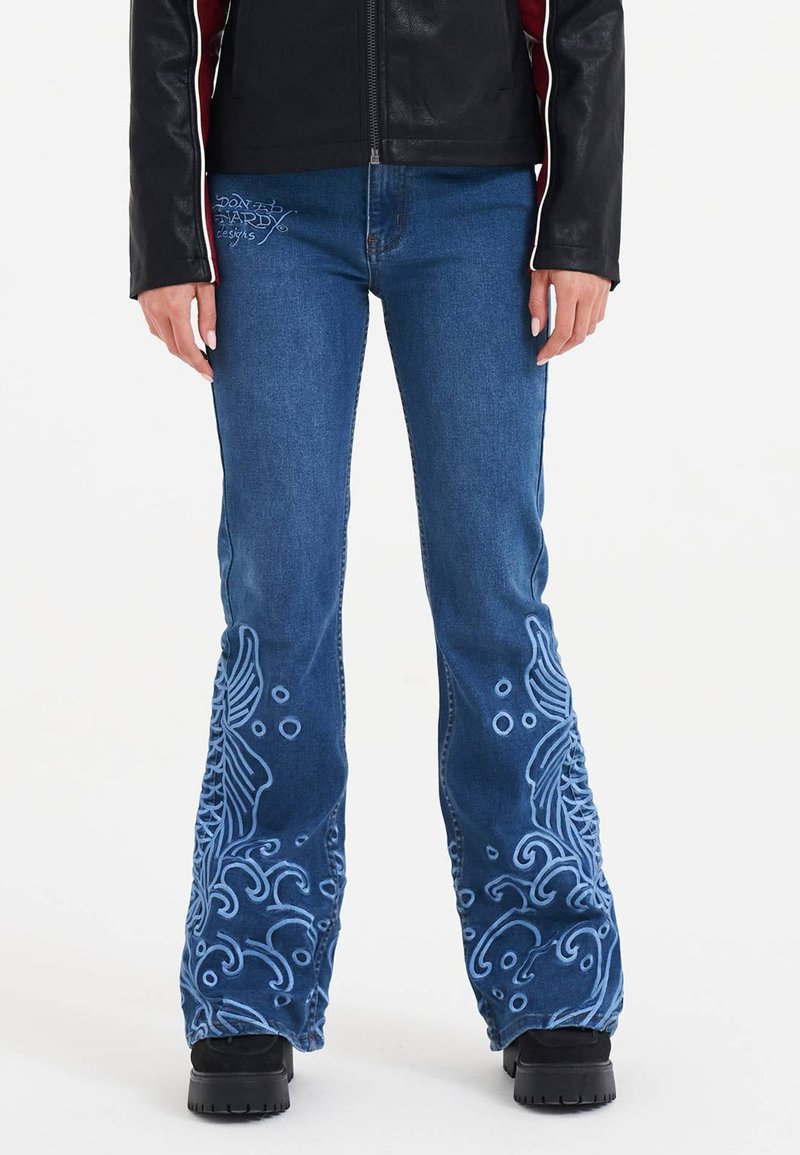 Bukser i medium blå denim med vidde, dekoreret med detaljeret lyseblåt broderi, der fremviser svirlende mønstre på benene, og en glat tekstur.