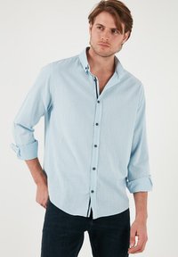 Camisa de botones de color azul claro, hecha de una tela texturizada, con cuello extendido, puños enrollados y botones oscuros, combinada con jeans oscuros.