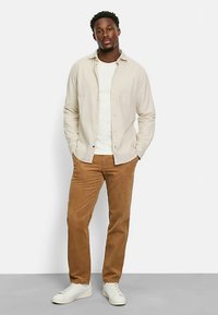 Beiges Hemd über einem weißen T-Shirt, kombiniert mit brauner Cordhose und weißen Turnschuhen, stehend vor einem neutralen Hintergrund.