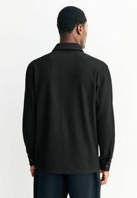 Melna garām piedurknēm krekls ar vertikāli ribotu tekstūru, ar klasisku apkakli un pogātām piedurknēm. Aizmugurē redzama brīva piegriezuma dizains.