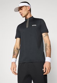 Svart kortärmad polotröja med dragkedja i kragen, med en vit "BOSS"-logotyp. Bärs med svarta byxor och vita handledband, som visar tatueringsdetaljer.