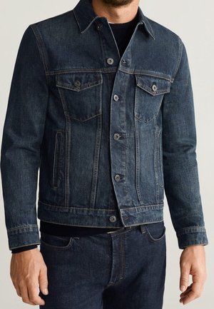 Mann trägt eine dunkelblaue Jeansjacke über einem schwarzen Hemd und passende dunkelblaue Jeans, steht entspannt mit locker hängenden Armen.