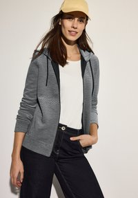 Felpa grigia con zip e cappuccio, con fodera nera a contrasto, indossata sopra una maglietta bianca, abbinata a jeans scuri e un cappello giallo chiaro.