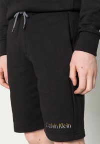 Pantaloni corti neri in cotone con vita elasticizzata e stringa. Presentano il logo ricamato "Calvin Klein" in giallo e bianco.