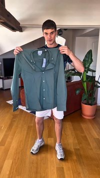 Cămașă verde cu mâneci lungi, cu nasturi, buzunar pe piept și guler. Materialul pare neted. Etichetă atașată, ținută de o persoană într-un living.