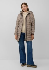 Abrigo acolchado beige con capucha, con un patr�ón acolchado y cremalleras negras, combinado con un suéter crema y vaqueros de pierna ancha azules.