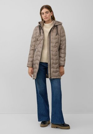 Beige gewatteerde jas met een capuchon, gekenmerkt door een gewatteerd patroon en zwarte ritsen, gecombineerd met een crèmekleurige trui en blauwe wijde jeans.