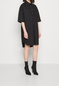 Czarna, oversize sukienka koszulowa z zapięciem na guziki z przodu, krótkimi rękawami i zaokrąglonym dołem. Połączona z teksturowanymi czarnymi botkami za kostkę.