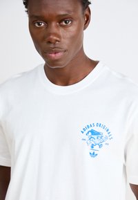 Λευκό βαμβακερό t-shirt με μπλε καρτούν και κείμενο "ADIDAS ORIGINALS" στο στήθος. Κοντά μανίκια, σχέδιο με στρογγυλή λαιμόκοψη.