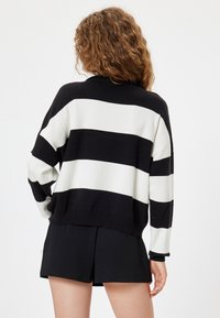 Gestreifter Pullover in Schwarz und Weiß mit lockerer Passform, langen Ärmeln und geripptem Saum. Getragen über schwarzen Shorts, was einen lässigen Stil zeigt.