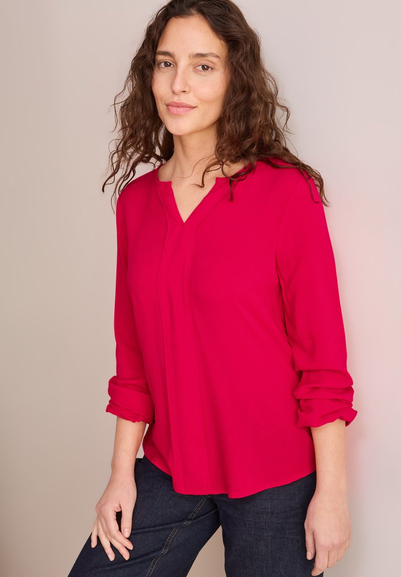Fuchsia långärmad blus med V-ringning och vikta ärmslut, i kombination med mörka denimjeans. Slät textur med subtila vertikala sömmar.