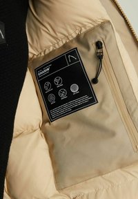 Wnętrze beżowej kurtki puchowej z czarną metką zawierającą instrukcje pielęgnacji oraz symbole odporności na wodę i wiatr, oddychalności i ciepła.
